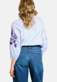 Femme blonde portant une blouse bleu rayé avec une broderie florale violette sur les manches et un jean taille haute bleu, vue de dos.