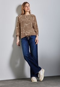Brun mönstrad tröja med ballongsärmar, stajlad med mörkblå vida jeans och krämfärgade sneakers. Mot en neutral bakgrund.