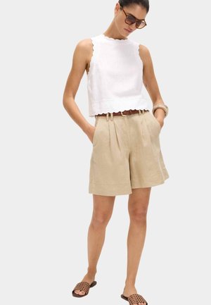 Frau trägt ein weißes ärmelloses Top, beige plissierte High-Waist-Shorts mit Gürtel, braune Sandalen, Sonnenbrille und Armband, Hände in den Taschen.