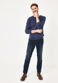 Marineblauer Rundhalsausschnitt-Sweatshirt, dunkelblaue Jeans und braune Schnürstiefel. Das Model steht mit verschränkten Armen und lächelt vor einem schlichten Hintergrund.
