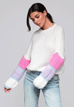 Pull en tricot blanc avec des manches rayées roses et bleues, col rond et coupe ample. Associé à un jean en denim délavé.