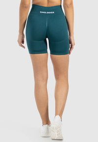 Vrouw in teal sportshorts en witte sneakers, gezien van achteren. De shorts hebben de merknaam "SMILODOX" op de tailleband.