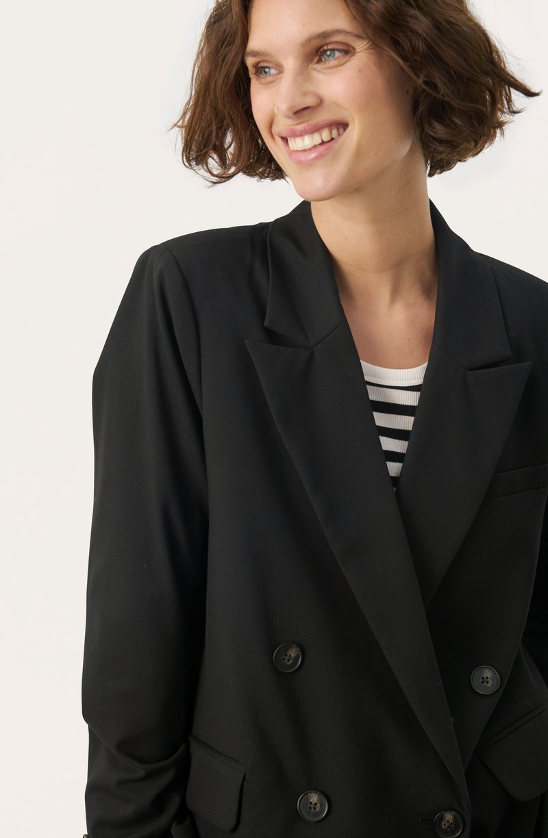 Schwarze, doppelte Blazerjacke mit Revers, großen Knöpfen und Taschen, über einem schwarz-weiß gestreiften Oberteil getragen.