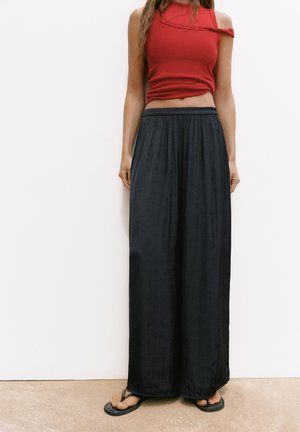 Femme portant un haut rouge sans manches, une jupe maxi noire ample et des tongs noires, debout contre un mur blanc.