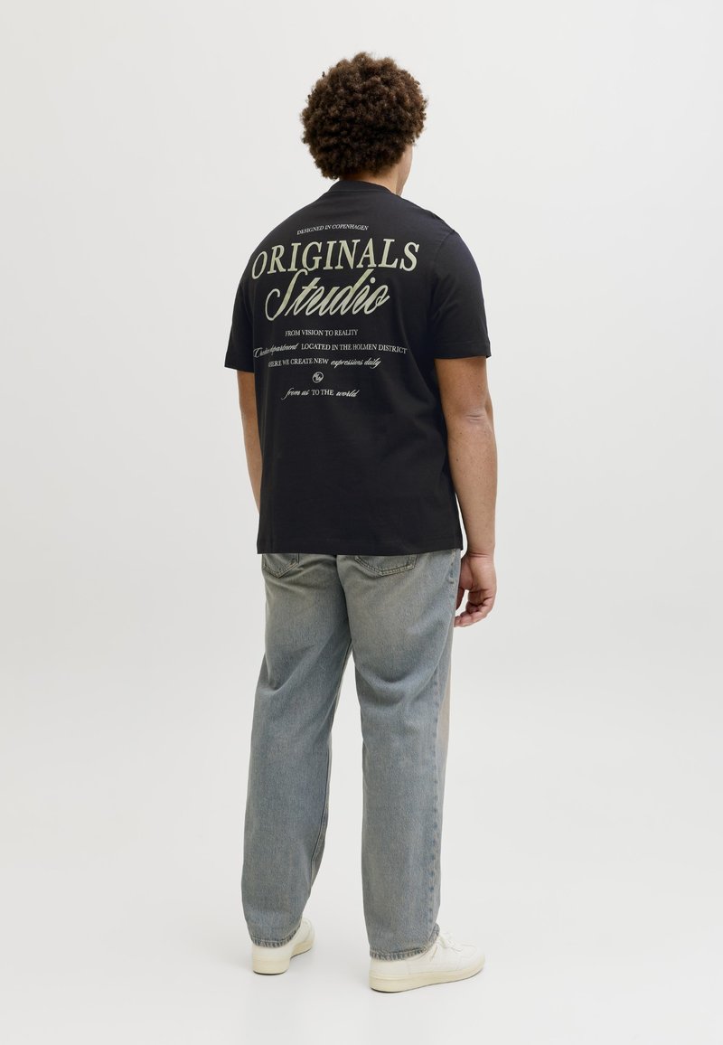 Göndör hajú férfi fekete "Originals Studio" feliratú pólóban, világoskék farmert és fehér tornacipőt viselve, egy egyszerű háttér előtt háttal áll.