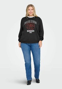 Mujer vestida con sudadera negra con una rosa roja y texto, jeans azules y botas negras, de pie contra un fondo claro y liso.
