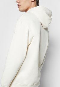 Sweat-shirt à capuche de couleur crème avec une coupe décontractée, tissu texturé, poignets côtelés et couture plate le long du dos.