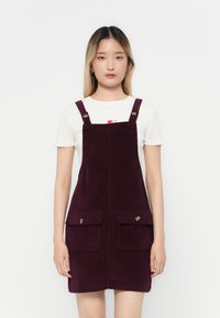Robe salopette en velours côtelé violet avec deux poches avant, bretelles réglables et boutons métalliques. Portée sur une chemise blanche avec des accents rouges.