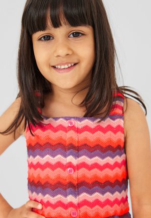 Jong meisje met donker haar, gekleed in een kleurrijke mouwloze top met zigzagpatroon, glimlachend met hand op de heup tegen een effen achtergrond.