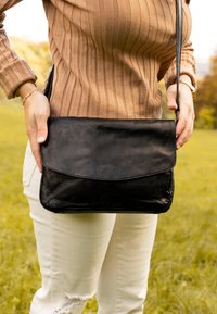 Schwarze Leder-Umhängetasche mit Klappenverschluss, glatter Textur, rechteckiger Form und verstellbarem Gurt, gehalten vor einer Person.