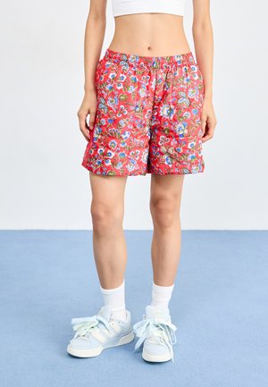 Shorts roșii cu flori, cu talie elastică, având modele de flori albastre și verzi. Asortați cu adidași albaștri deschiși și șosete albe.