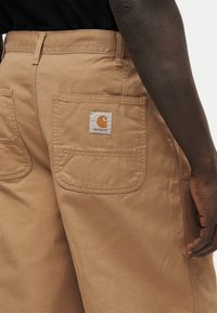 Κοντινό πλάνο ενός ατόμου που φορά παντελόνια Carhartt σε ανοικτή καφέ απόχρωση, με ορατή πίσω τσέπη και το έμβλημα της Carhartt, ενώ το χέρι βρίσκεται στο πλάι.