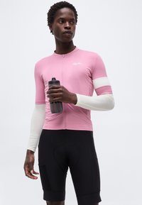 Person trägt ein pinkes Radtrikot über einer weißen langärmligen Basisschicht und schwarzen Radhosen und hält eine schwarze Wasserflasche.