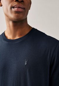 Marineblaues Baumwoll-T-Shirt mit rundem Halsausschnitt und einem dezenten, auf der linken Brust eingeprägten Logo. Glatte Textur, lässige Passform.