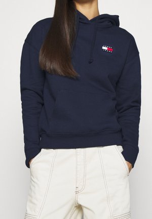 Persoon die een navyblauwe Tommy Jeans hoodie met voorzak draagt en witte broek met zichtbare stiksels, handen in de zakken, lang steil haar.
