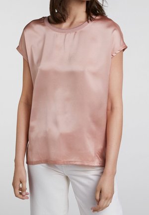 Femme portant un chemisier en satin rose brillant à manches courtes et un pantalon blanc, debout les bras détendus le long du corps.