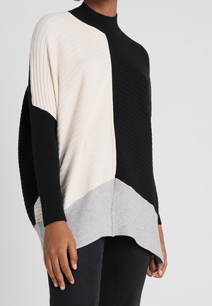 Femme portant un pull côtelé à blocs de couleurs noir, blanc et gris, associé à un pantalon noir, debout devant un fond uni.