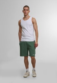 Weißes Baumwoll-Tanktop mit breiten Trägern, kombiniert mit grünen shorts bis zum Knie, die ein dezentes Logo zeigen. Neutrale Sneaker runden den Look ab.