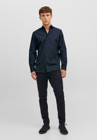 Tamno plava košulja dugih rukava, slim fit kroja, izrađena od teksturiranog materijala, uparena s tamnim hlačama i crnim cipelama. Minimalistički dizajn, bez uzoraka.