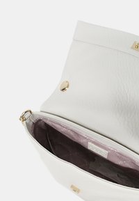 Hvid læderhåndtaske med tekstureret overflade og guldhardware. Magnetlukningen afslører et mauve-farvet indre med et mærke.