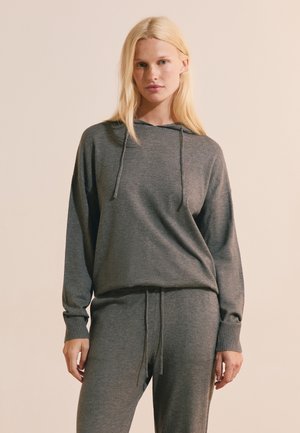 Femme blonde portant un sweat à capuche gris et un pantalon de jogging gris assorti, debout devant un fond beige uni.