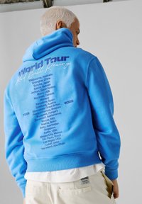 Άτομο με κοντά λευκά μαλλιά που φοράει φωτεινό μπλε φούτερ με κουκούλα, με καταχωρισμένες τις παγκόσμιες τοποθεσίες και τα έτη αγώνων τυπωμένα στην πλάτη.