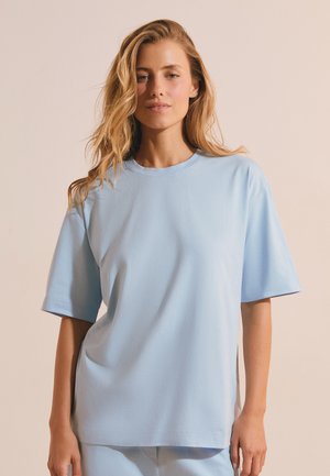 Jeune femme aux longs cheveux blonds portant un t-shirt oversize bleu clair à manches courtes sur un fond beige uni.
