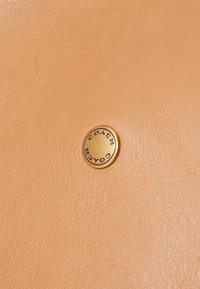 Tanleren oppervlak met een gladde textuur, voorzien van een ronde gouden metalen sierdeel met het gegraveerde merk "COACH".