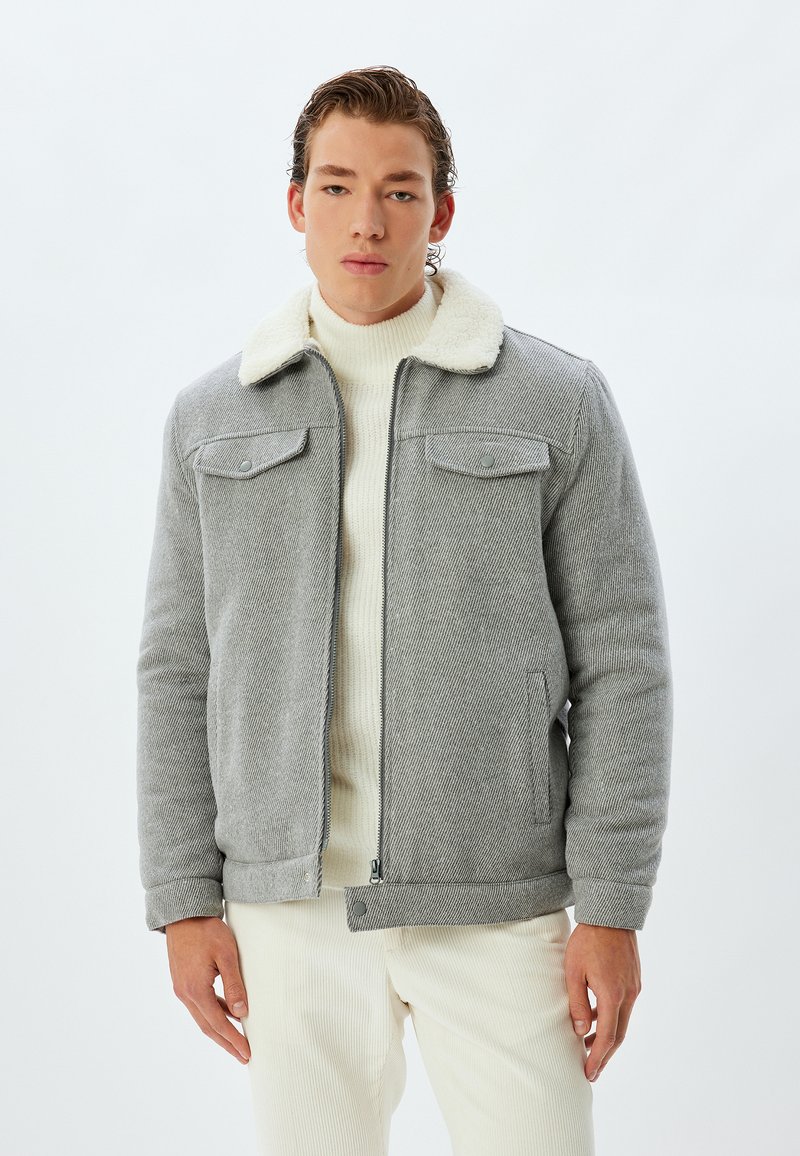 Grå, struktureret jakke med shearling-krave, lynlåslukning og frontlommer, lagdelt over en cremefarvet turtleneck. Varmt, afslappet design.