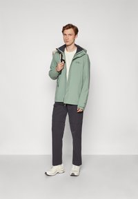 Hellgrüne wasserdichte Jacke mit grauem Kapuze, Reißverschluss und Logo auf der Brust. Getragen mit dunklen Hosen und weißen Sneakers.