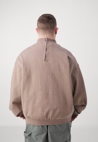 Uomo di spalle che indossa un felpa oversize beige con zip sul retro e pantaloni cargo grigi, contro uno sfondo bianco semplice.