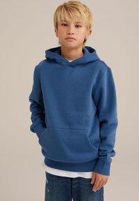 Sudadera con capucha azul de material suave, con un bolsillo frontal y puños de canalé, llevada sobre una camiseta blanca y jeans de mezclilla desgastados.