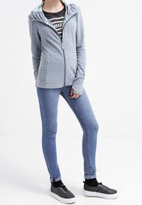 Giacca grigia con zip, con interno azzurro chiaro, indossata sopra una maglietta a righe nere e jeans skinny blu, abbinata a sneakers nere.