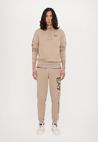 Beige hettegenser med lomme foran og detaljerte svarte grafikker på ertene; matchende beige joggebukser med logotrykk på benet; hvite sneakers.