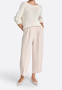 Suéter de punto crema con mangas holgadas combinado con pantalones plisados de color beige claro. El modelo lleva zapatos de punta de color claro.