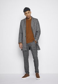 Manteau à carreaux gris, pull en laine brun, pantalon à carreaux et baskets brunes. Les accessoires comprennent une casquette sombre. Textures lisses, coupe ajustée.