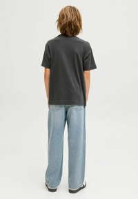 Persona con cabello hasta los hombros que lleva puesta una camiseta de manga corta gris oscuro, unos vaqueros anchos de color azul claro y zapatos en blanco y negro, de espaldas.