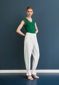 Green lace-trimmed bodysuit paired with white high-waisted trousers, featuring pleats and knotted cuffs, complemented by strappy beige sandals.