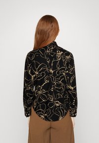 Camicia nera con stampa floreale dorata, maniche lunghe e orlo arrotondato. Dettaglio con piega sul retro e polsini con bottoni. Tessuto di texture liscia.