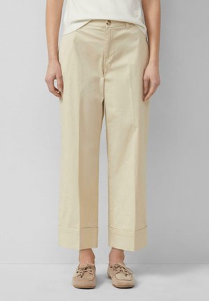 Person trägt beige, weit geschnittene, gekürzte Hosen mit Umschlägen, kombiniert mit beigen Mokassin-Schuhen und einem weißen Oberteil, steht vor einem schlichten Hintergrund.