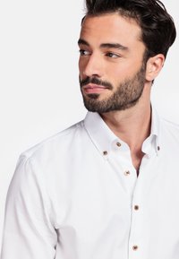 Witte knop-up shirt met een klassieke kraag, bruine knoopaccenten en een gladde, textuurstof. Close-up van het shirt met de schouder van het model.