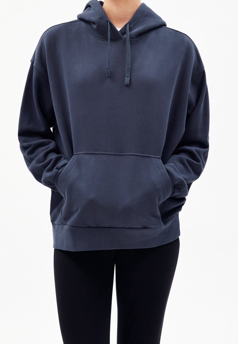 Personne portant un sweat à capuche bleu foncé avec une poche kangourou à l'avant et un pantalon noir, debout les mains dans les poches sur un fond uni.