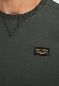 Felpa di cotone verde scuro con colletto tondo a coste, caratterizzata da un patch del marchio ricamato in nero e oro sul lato sinistro.