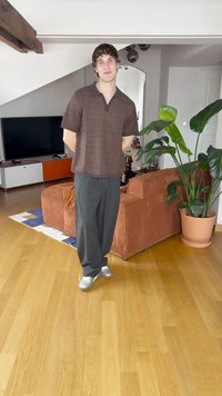 Camisa polo castanha de mangas curtas com um padrão texturizado, calças pretas relaxadas e ténis prateados, contra um fundo de sala de estar moderna.