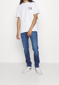 Vit bomulls t-shirt med flerfärgad "TOMMY" logotyp på bröstet, kombinerad med blå denimjeans och vita sneakers.