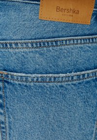 Ljust blå denimjeans med en bakficka, med en brun läderetikett sydd vid midjan med texten "Bershka EST. 1998."
