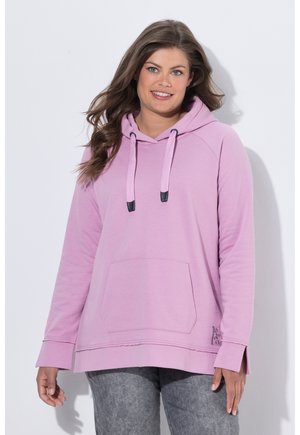 Frau mit langen braunen Haaren, die einen hellvioletten Hoodie mit Fronttasche und graue Jeans trägt, lächelnd und vor einem weißen Hintergrund steht.