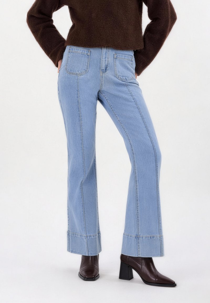 Γυναίκα που φοράει τζιν ελαφρώς μπλε με ίσια μπατζάκια και μεγάλες μπροστινές τσέπες, συνδυασμένα με σκούρα καφέ μποτάκια με τακούνια και σκούρο καφέ πουλόβερ.