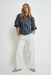 Denimbluse mit Puffärmeln, Knopfverschluss, dunkelgraue Farbe; kombiniert mit weißen, weit geschnittenen Hose und beigen Ballerinas.