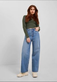 Πράσινη μπλούζα μακρύ μανίκι σε συνδυασμό με μπλε τζιν ψηλής μέσης και φαρδιάς εφαρμογής. Τα τζιν διαθέτουν κλασικό σχέδιο πέντε τσεπών και ανοιχτό χρώμα.